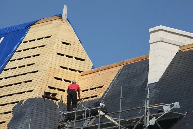 Couvreur sur un échafaudage en train de poser des ardoises devant une façade en bois en construction.