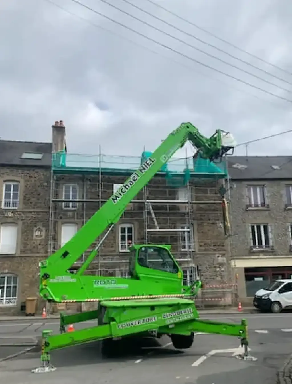 Engin de levage vert devant une maison en pierre avec échafaudage, sous un ciel clair