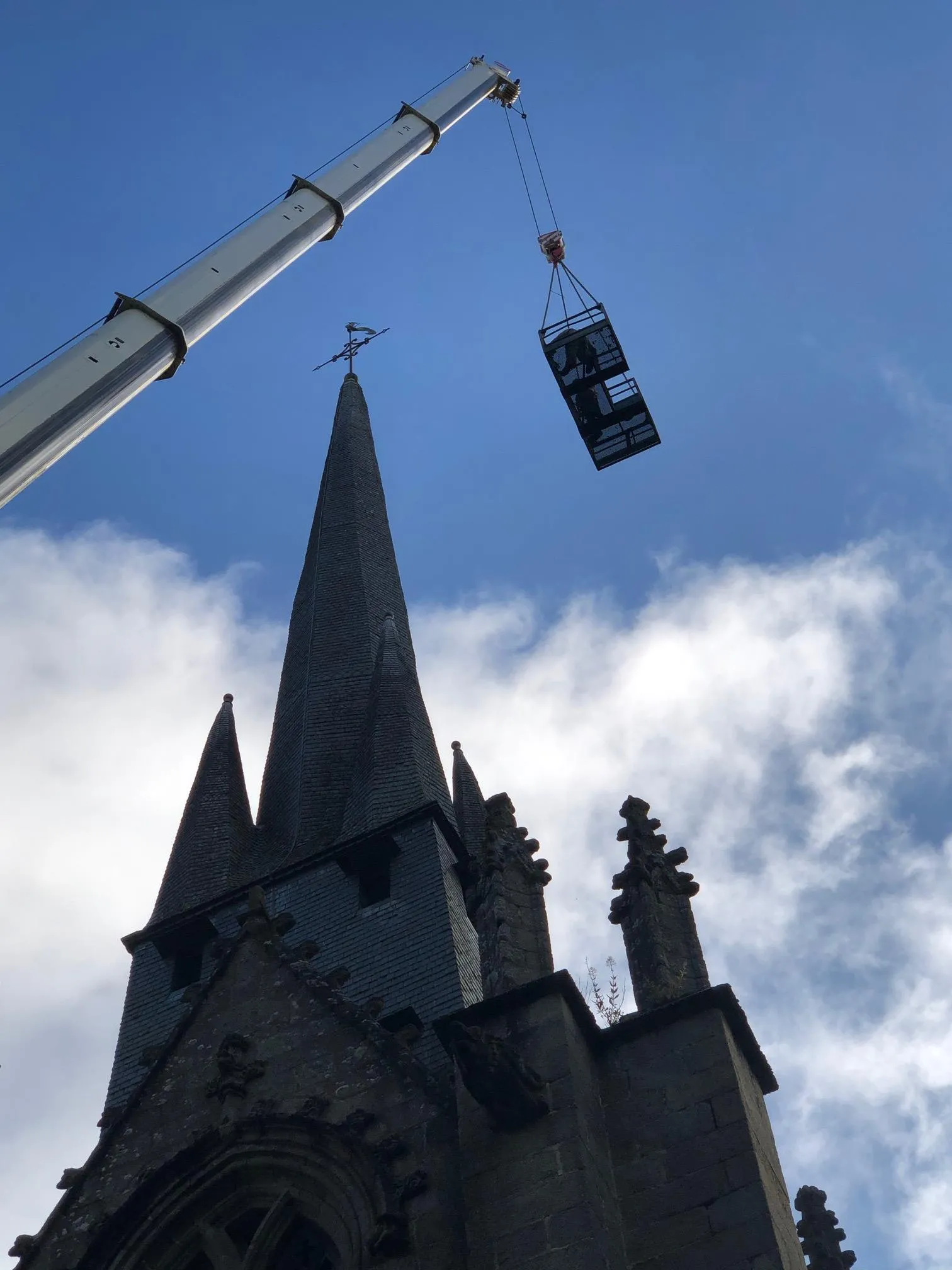 Flèche en ardoise d'un clocher en pierre, avec une nacelle suspendue à une grue