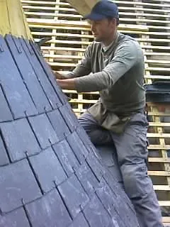 Couvreur posant de l'ardoise sur un arêtier, avec un lattage visible derrière lui