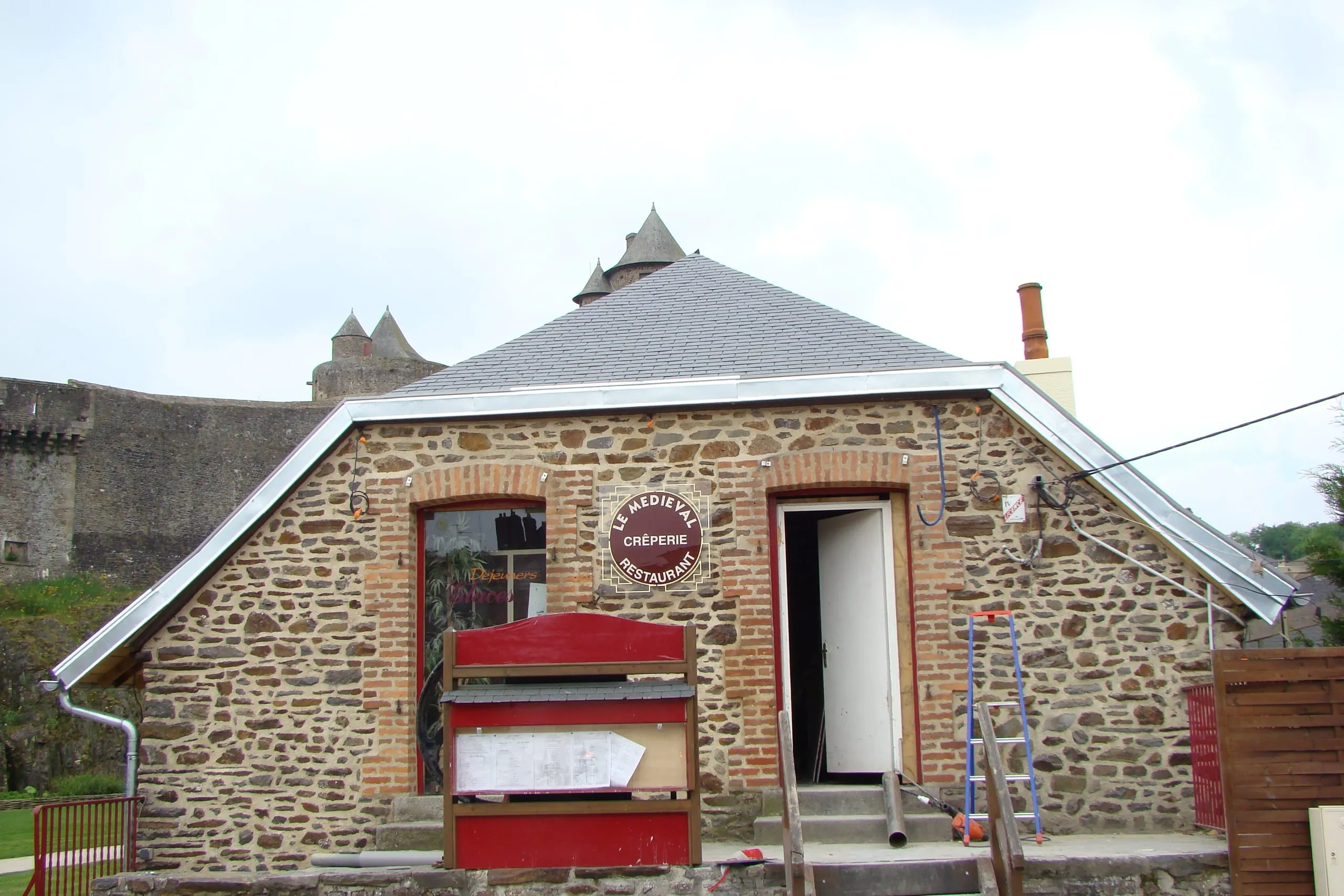 Façade en pierre et brique d'une crêperie avec une toiture en ardoise bordée de zinc, devant les murs d'un château