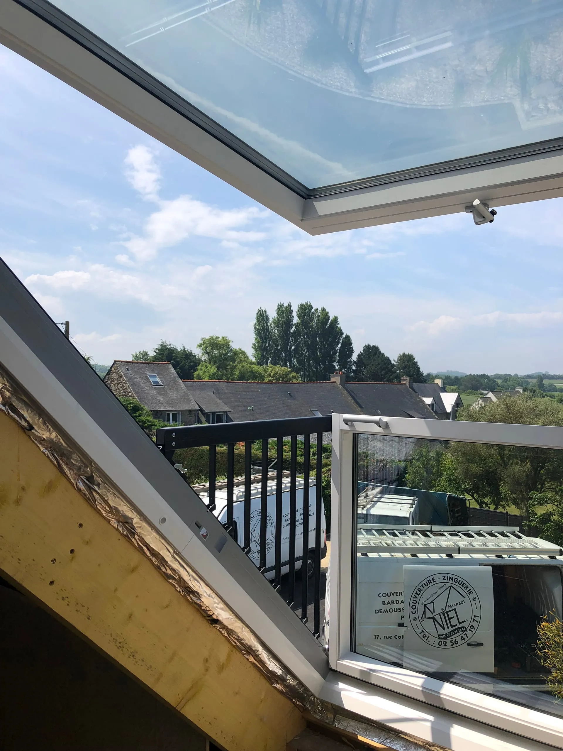 Fenêtre de toit à ouverture en balcon ouverte, avec vue sur des toits en ardoise et sur un camion de l'entreprise