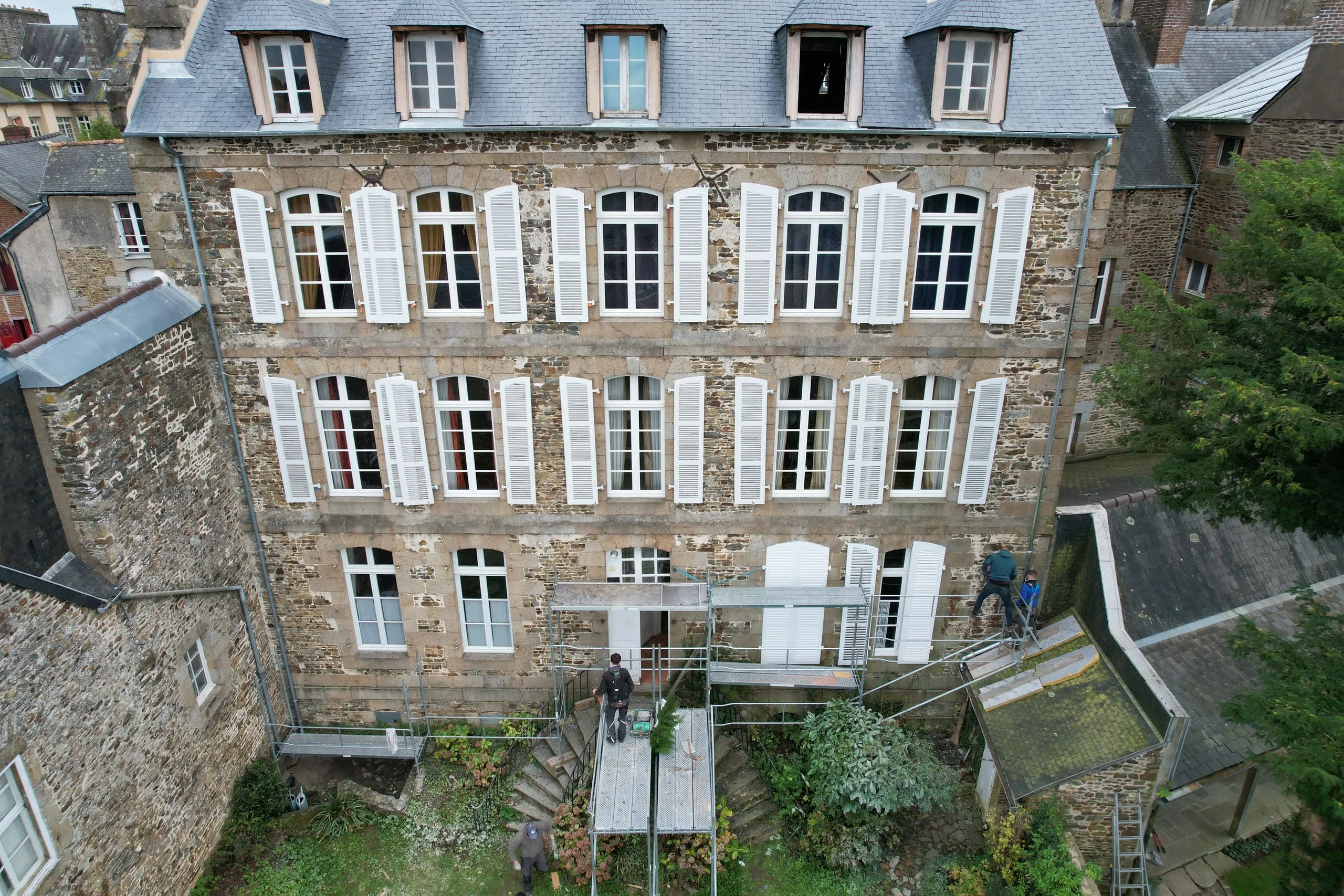 Façade en pierre à volets blancs d'un grand bâtiment ancien, vue du dessus, avec un échafaudage et des ouvriers en bas
