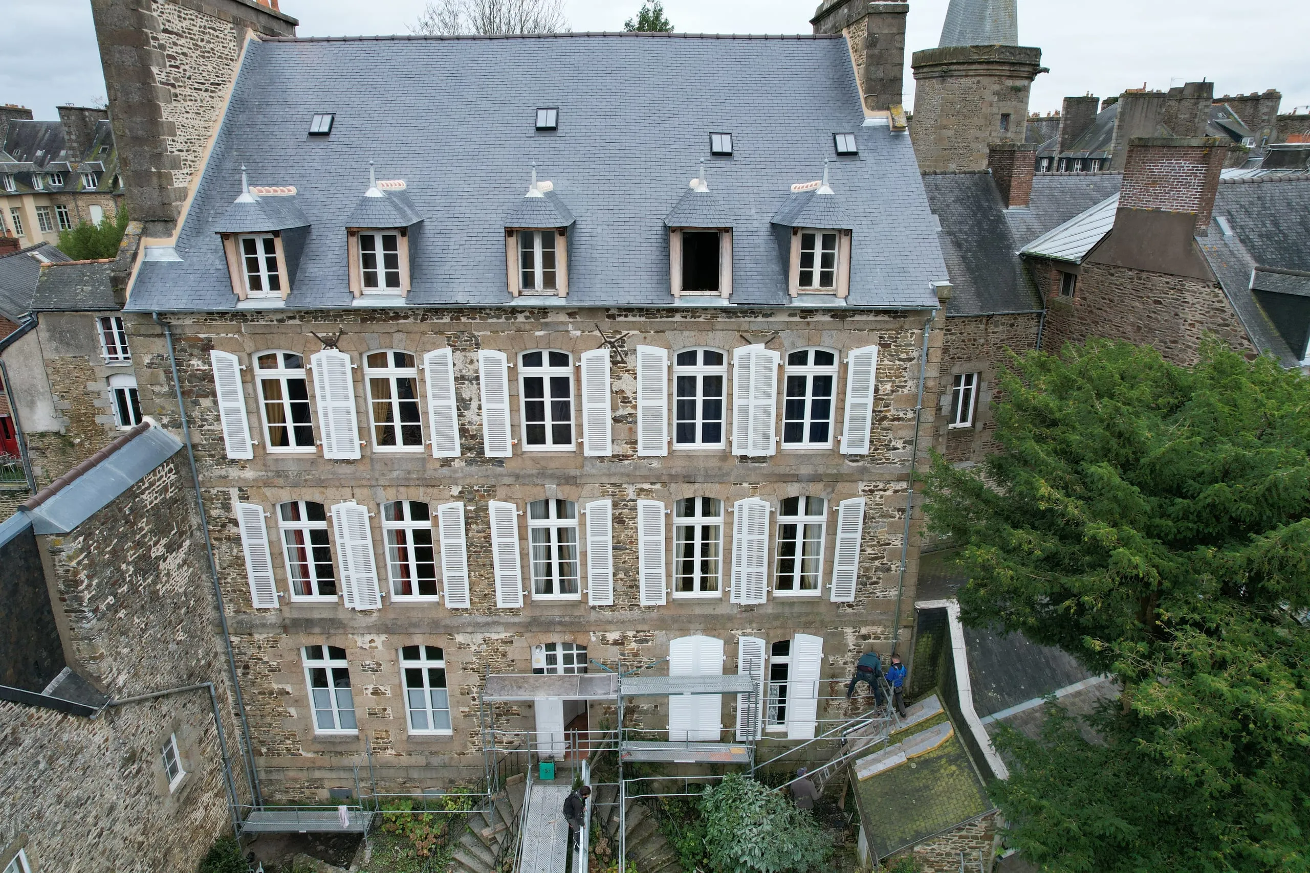 Façade en pierre à trois niveaux de fenêtres et volets blancs, sous un toit en ardoise à cinq lucarnes et quatre fenêtres de toit