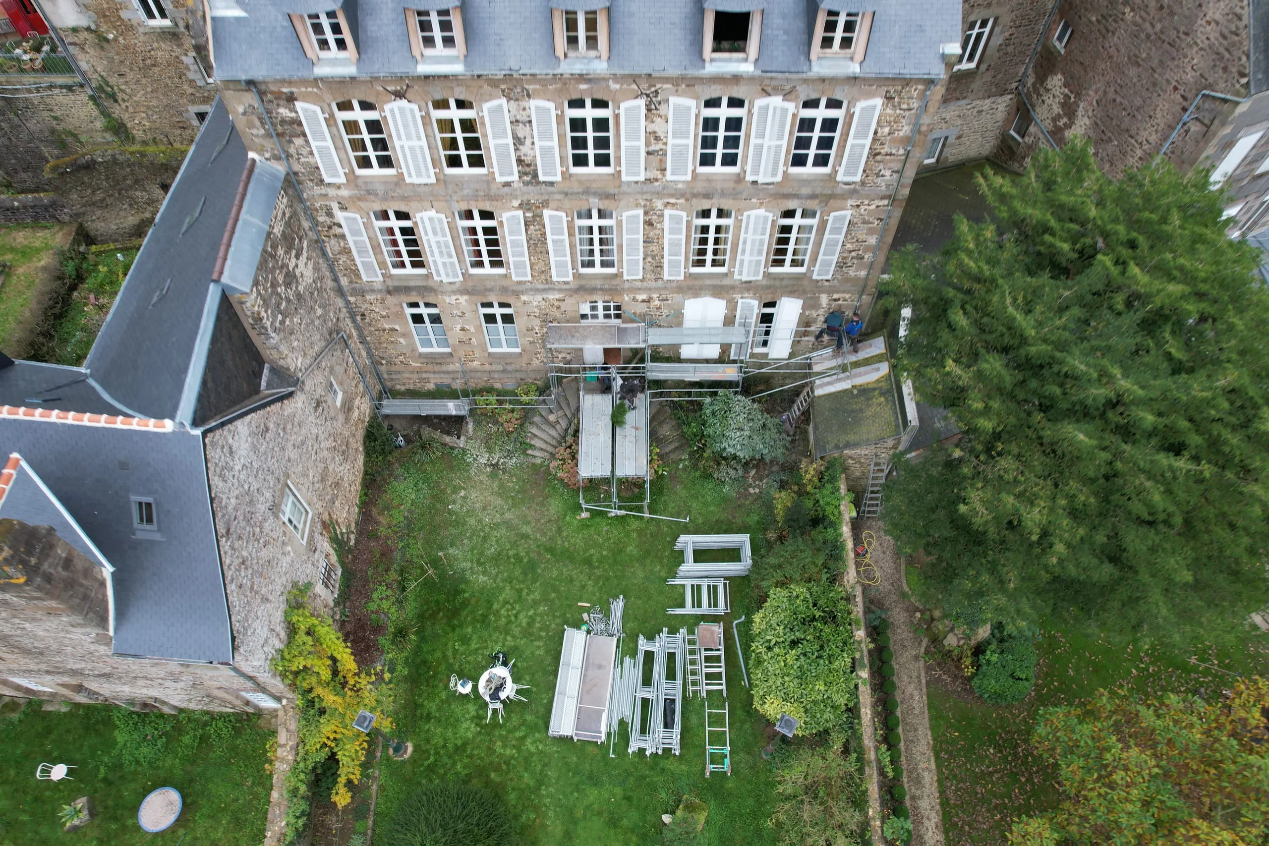 Vue aérienne d'un grand bâtiment en pierre à volets blancs, avec un échafaudage et du matériel stocké dans le jardin